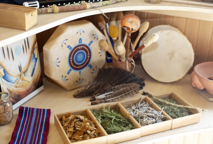 A wooden shelf displays various objects including a framed painting, two drums, a quiver of feathers, and wooden maracas. In the foreground are four compartments containing different dried herbs. A colorful woven cloth and a small jar are also visible.