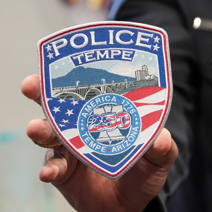 A person holds a Tempe Police badge featuring the word "POLICE" above the city name "TEMPE," with a background of mountains, the American flag, and a cityscape. The badge also includes "America 1776," "250," and "Tempe, Arizona" in the design.