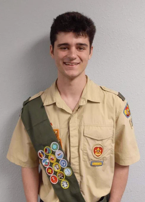 A smiling young man in a tan shirt and green sash with various badges stands against a white background.