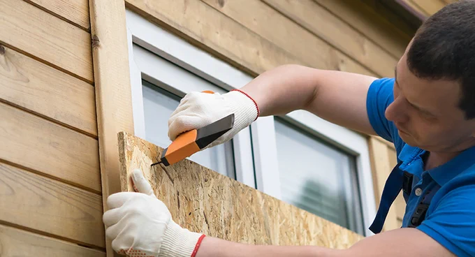 Person wearing white gloves using a utility knife to cut plywood against a wooden house exterior near a window.