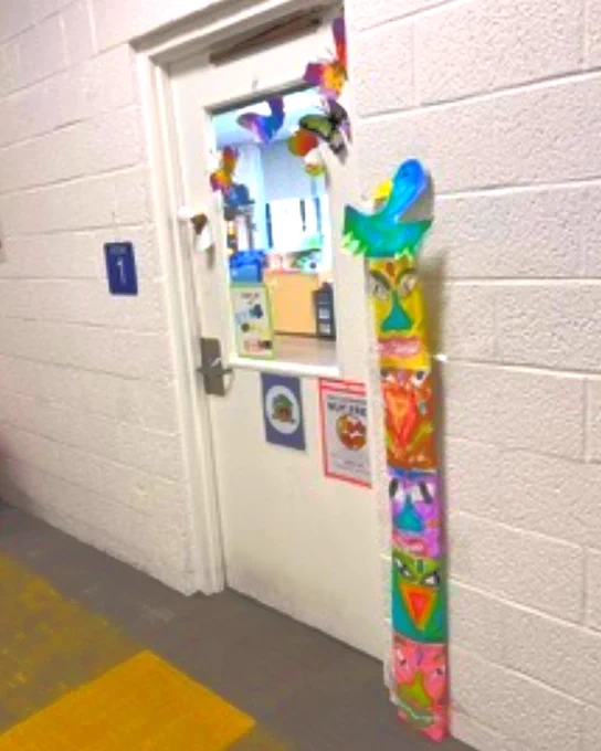 Classroom door decorated with colorful paper crafts and a large blue bird cutout on a white brick wall.