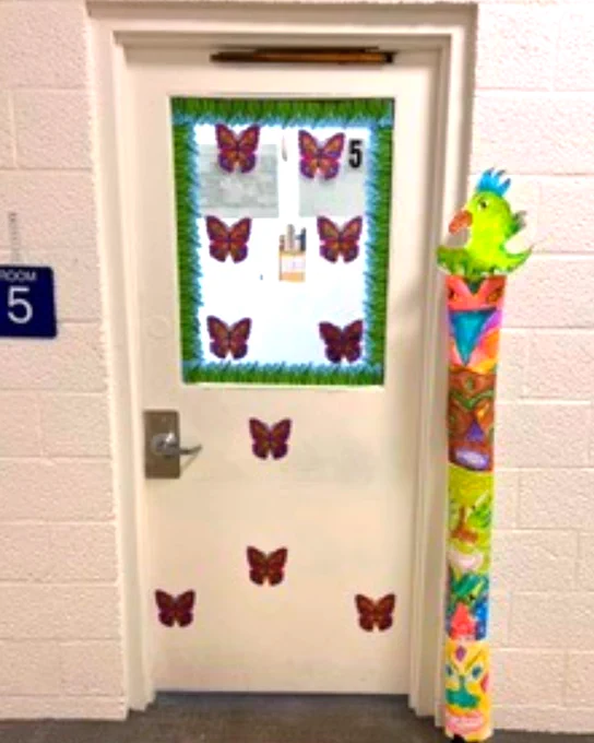 White door decorated with multiple butterfly cutouts and a green tinsel border, labeled as Room 5 in a hallway.