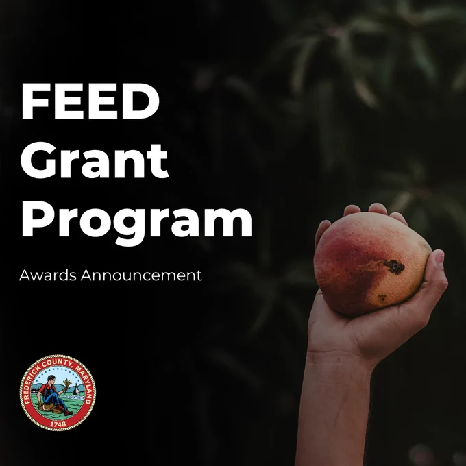 A promotional graphic features a dark, softly blurred background with the silhouette of foliage or tree branches. On the left side, large bold white text reads “FEED Grant Program,” and directly beneath it in smaller white text are the words “Awards Announcement.” In the bottom-left corner, the Frederick County, Maryland seal is displayed, showing an illustrated scene with a person seated near water and the year 1748. On the right side of the graphic, a hand extends upward holding a ripe peach 