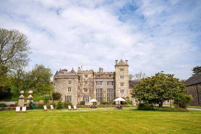 A historic stone manor with towers and large windows under a partly cloudy sky. The lush green lawn features deck chairs and small tables with umbrellas.