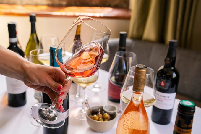 A person holding a tilted glass of rosé wine over a table set with various wine bottles, glasses, and a bowl of olives, conveying a social and elegant atmosphere.
