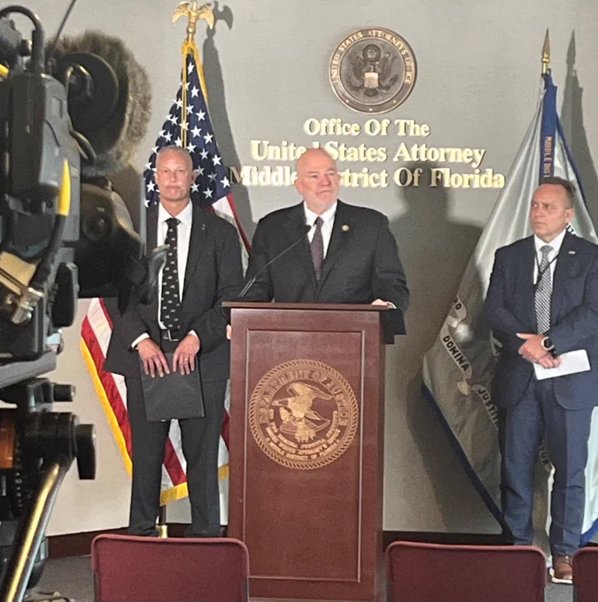 USA Kehoe at podium flanked by federal officials.  DOJ seal and flags in background.