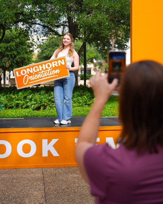 Mom takes a photo of her daughter holding a sign to celebrate UT Austin Orientation. 
