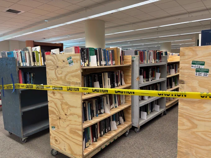 Bookshelves on wheels in a library. Yellow caution tape is draped across the shelves, indicating restricted access. Various books are visible on the shelves.