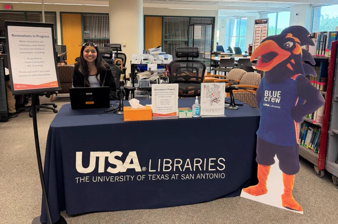 This image shows a person sitting at a desk covered with a UTSA Libraries tablecloth, in an academic setting. A sign on the table reads "Renovations in Progress," detailing areas being improved. Next to the table is a standee of Rowdy the Roadrunner, the mascot for the University of Texas at San Antonio. Bookshelves and computers are visible in the background.