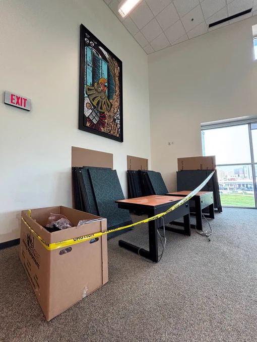A room with a long, L-shaped table partially surrounded by caution tape. Stacked chairs are pushed against the wall. There is a large decorative wall art piece and an exit sign nearby. A cardboard box sits on the carpeted floor. A window in the background shows a grassy area outside.