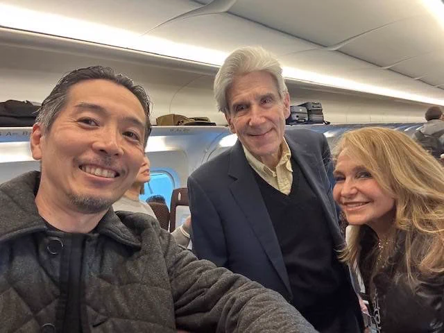 Three casually dressed people posing for a selfie group photo in a plane cabin. The interior features overhead storage compartments and a window on the left side.
