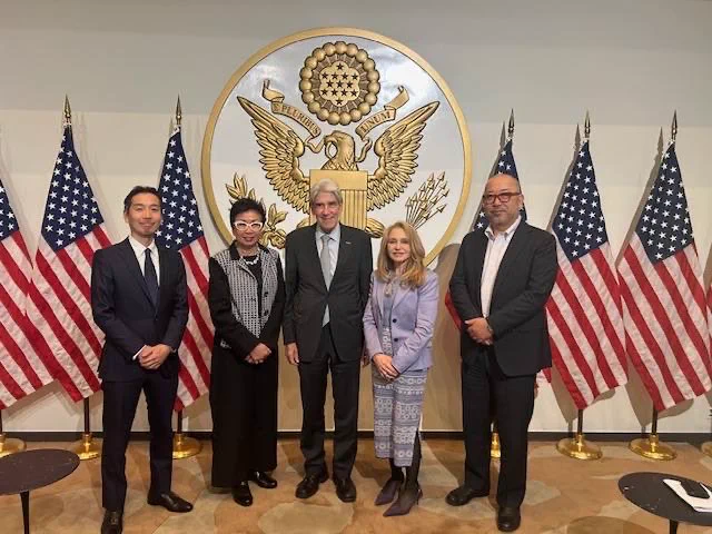 Five people in formal attire stand in front of a large circular emblem featuring the Great Seal of the United States at the US Embassy in Tokyo. The seal is flanked by multiple American flags arranged symmetrically.