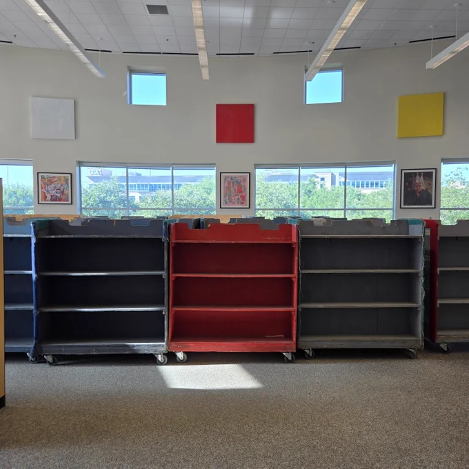 Empty red and black bookshelves on wheels are arranged in a line inside a library. Large square artworks hang above them, and windows in the background reveal a view of trees and a building.