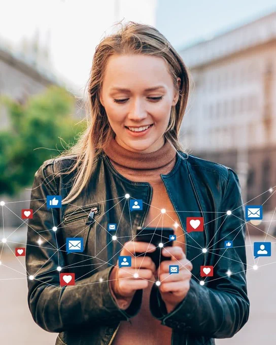 Person using a smartphone with social media icons like hearts, thumbs up, and envelopes floating around them, outdoors.
