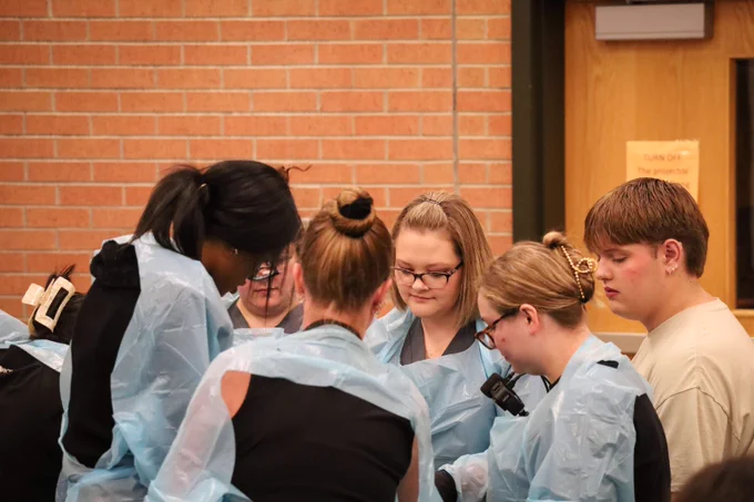 Group of people wearing light blue protective gowns gathered closely, engaged in a focused discussion against a brick wall background.
