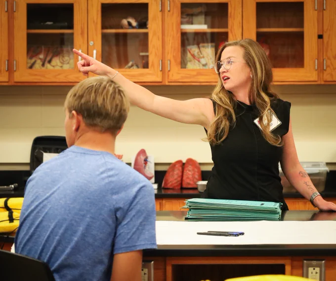 Teacher in a classroom pointing towards the cabinets while a student in a blue shirt listens attentively.