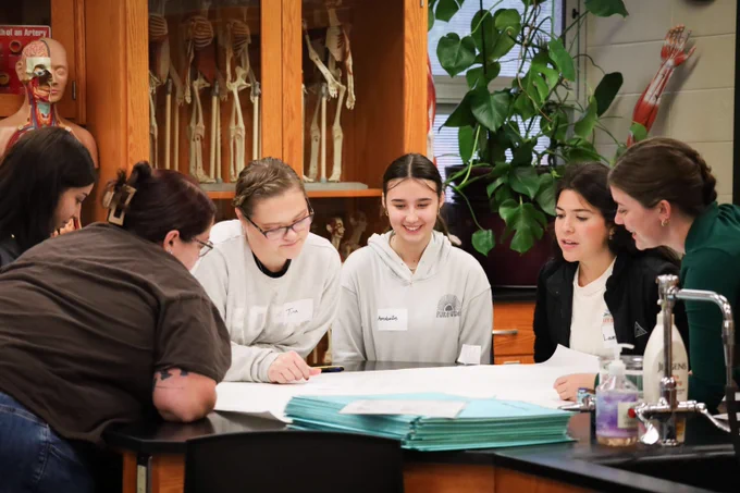 A group of students gathered around a lab table, engaged in a collaborative discussion with anatomical skeleton models in the background.