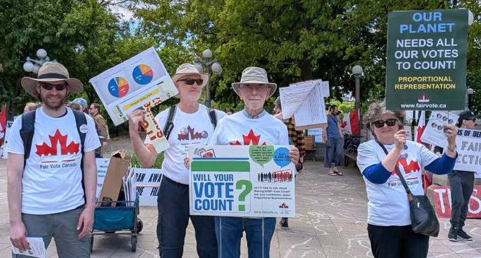 A group of five Fair Vote Canada volunteers stands together outdoors in a paved public square. They wear matching white t-shirts featuring a red stylized Canadian maple leaf and the words Fair Vote Canada. The volunteers hold up campaign materials, including a large white sign that reads Will Your Vote Count? next to a green box. Another volunteer holds a dark green sign that says Our Planet Needs All Our Votes to Count! Proportional Representation. In the background, green trees and park benches are visible under a bright sky.