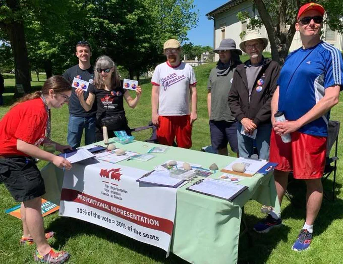 A group of volunteers gathers around a green-clothed campaign table on a grassy lawn under a clear blue sky. A white banner attached to the front of the table reads Proportional Representation 30% of the Vote = 30% of the Seats, alongside a Fair Vote Canada logo. Clipboards with sign-up sheets are neatly arranged on the table, held down by small rocks. Two volunteers hold up small informational pamphlets, while others stand behind the table interacting during a sunny outdoor community event.