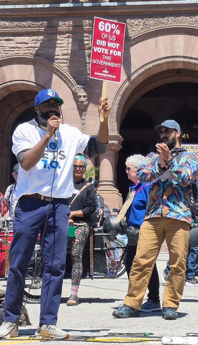 A speaker stands outdoors in front of a stone building with large Romanesque arches, addressing a crowd through a microphone. He wears a white t-shirt, dark trousers, and a blue baseball cap. In his left hand, he holds high a red rectangular sign that reads 60% of Us Did Not Vote for This Government! FairVote.ca. To his right, another man in a colourful patterned jacket and a grey cap stands clapping, while other demonstrators and a drum set are partially visible in the background.