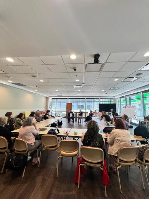 A wide shot of a large conference room where participants are seated along a U-shaped table setup, facing three speakers at the front.