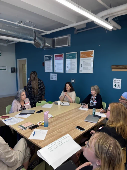 A group of people sitting around a large wooden table in a meeting room with a blue wall, engaged in a discussion with papers and notebooks open.