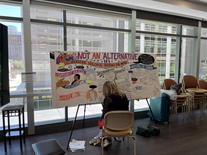 A person sits from behind, working on a large, colorful graphic recording board titled "NOT AN 'ALTERNATIVE'" in front of a large window.