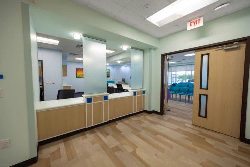 A modern medical office reception area with light green walls, wooden flooring, and a counter with privacy screens. An open door leads to a waiting room with teal chairs.
