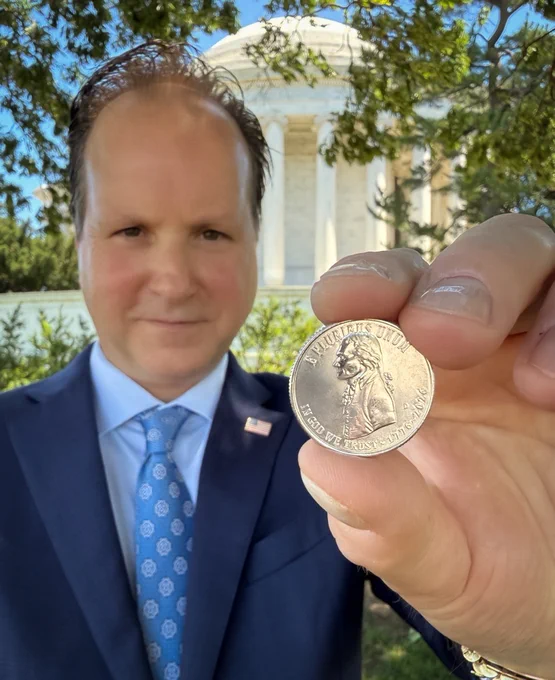 U.S. Mint Director Paul Hollis holds up a Quarter with Thomas Jefferson's profile. The background features the Jefferson Memorial surrounded by trees.