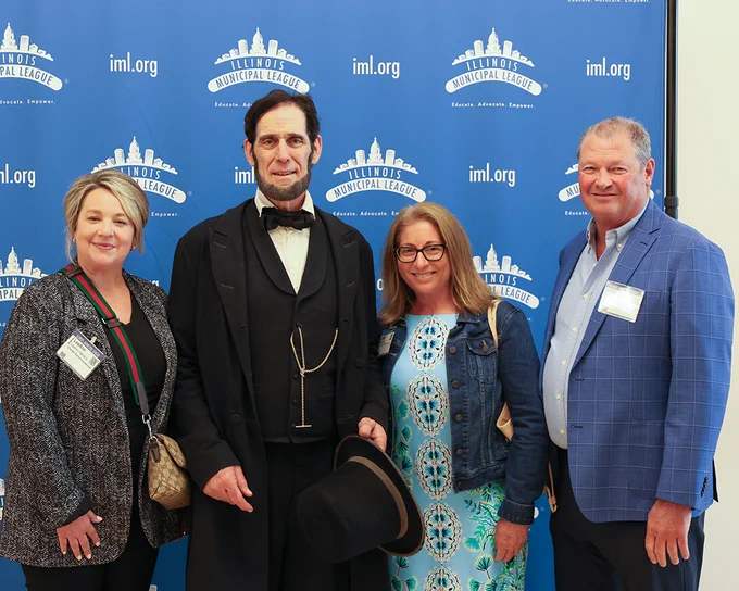 Four people standing in front of an Illinois Municipal League backdrop, one dressed as Abraham Lincoln.
