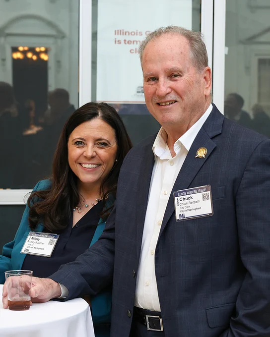 Two professionals wearing name badges stand by a table with a drink, attending an event in a formal setting.