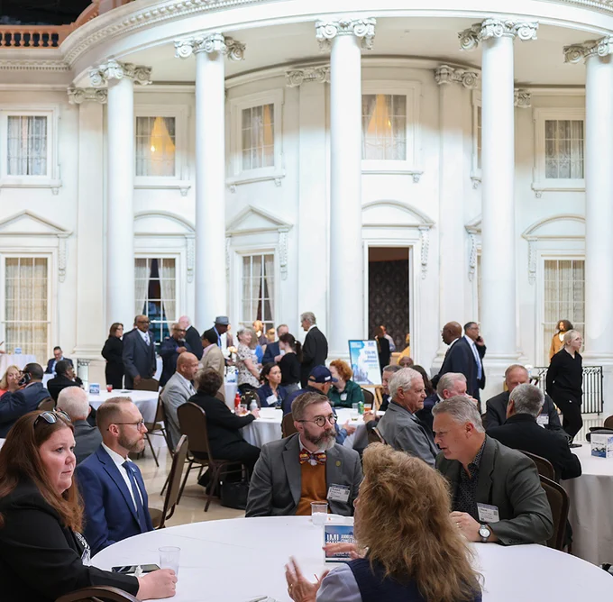 Guests engage in conversation at round tables during a formal event inside a grand hall with tall white columns and large windows.
