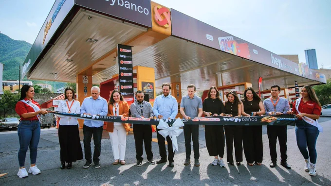 Un grupo de personas participa en una ceremonia de corte de cinta frente a una estación de servicio cubierta, con carteles de "heybanco" y "Super Cashback". Un gran lazo blanco adorna la cinta. Al fondo se ven montañas y edificios.
