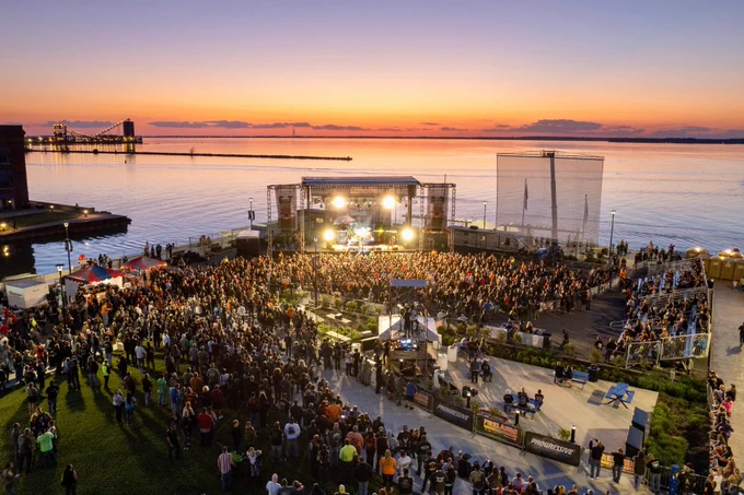 Outdoor concert at dusk with a band performing on a stage to a large crowd by the water.
