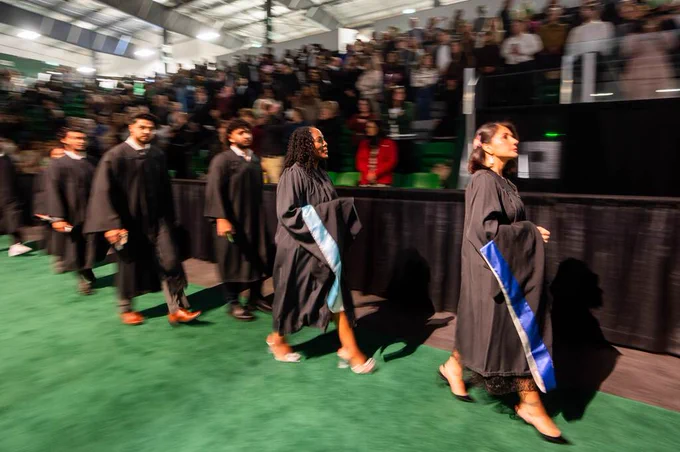 A group of graduates in caps and gowns walk in a procession on a green carpet during a graduation ceremony, with a large audience seated in the background.
