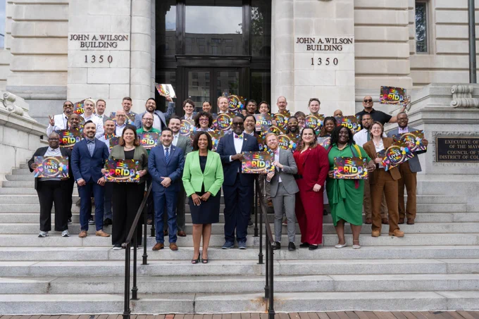 Mayor Bowser stands with individuals who identify as members of the LGBTQIA  community on the steps of the John A. Wilson Building. Some individuals are holding colorful signs that say "District of Pride."