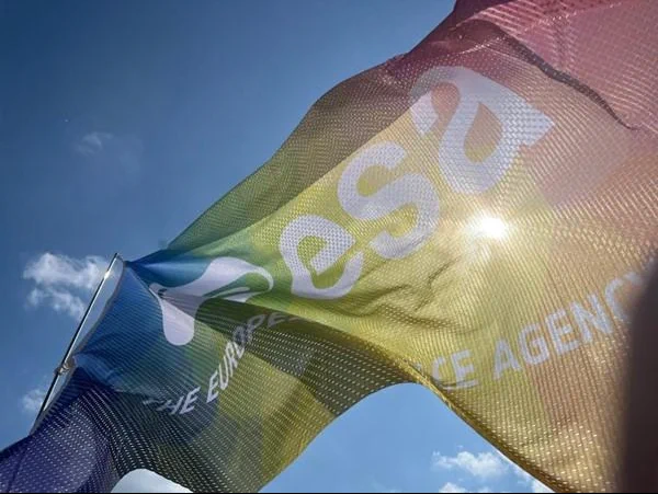 A rainbow‑coloured ESA (European Space Agency) flag waves in the wind against a bright blue sky with scattered clouds. Sunlight shines through the semi‑transparent fabric, making the ESA logo and text partially visible across the flowing material.