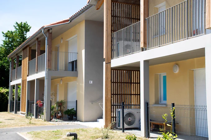 Façade de la résidence. Le bâtiment associe des murs enduits de couleur beige clair et des structures en bois (poteaux, pare-vue ajourés). Les logements du rez-de-chaussée possèdent de petits espaces extérieurs délimités par des clôtures grillagées.