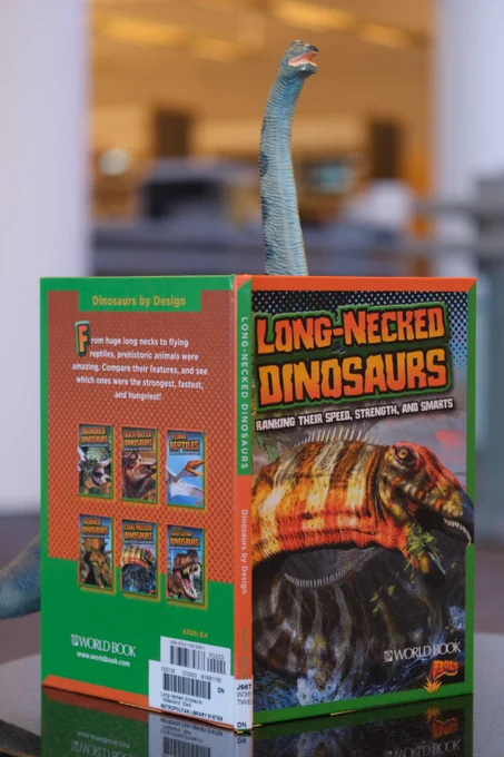 A toy long-necked dinosaur peeks over the top of an open book titled 'Long-Necked Dinosaurs' with colorful illustrations on the cover and back.