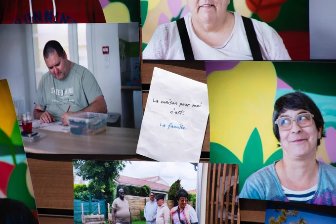 Un panneau d'affichage en bois sur lequel sont épinglées plusieurs photos de résidents dans leur quotidien (un homme qui dessine, un groupe dans un jardin, des portraits colorés). Au centre, un petit mot manuscrit indique : « La maison pour moi c'est : La famille. » 