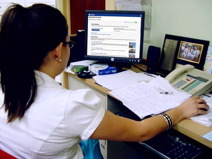 A person is sitting at a desk using a computer. The screen displays the "Seattle Services Portal." There are various papers and office supplies on the desk, including a telephone, tissue box, and framed group photograph.
