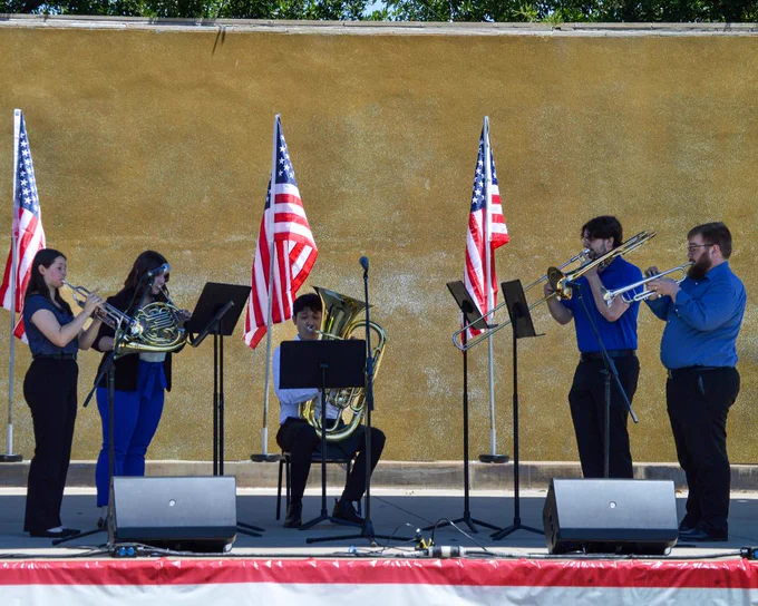 A brass quintet performs on an outdoor stage, decorated with three American flags.