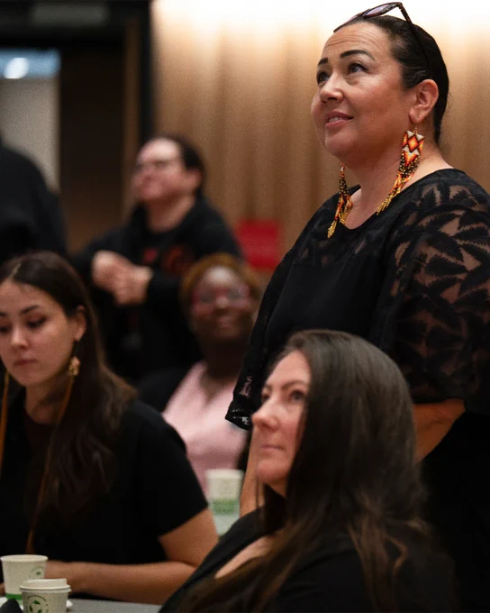 Alt text: A participant wearing beaded earrings stands and speaks during a group information session while other attendees listen and engage in discussion around the room.