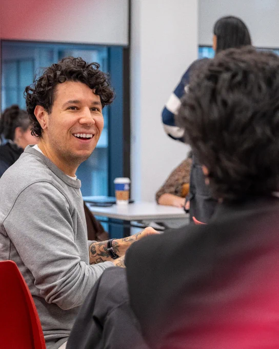 Alt text: A smiling participant sits at a table during a group discussion or information session, looking toward another person in the room. Other attendees are visible in the background of the bright meeting space.