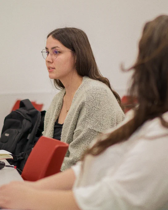 Alt text: A participant wearing glasses and a light cardigan listens attentively during a small-group discussion or information session, seated at a table with other attendees.