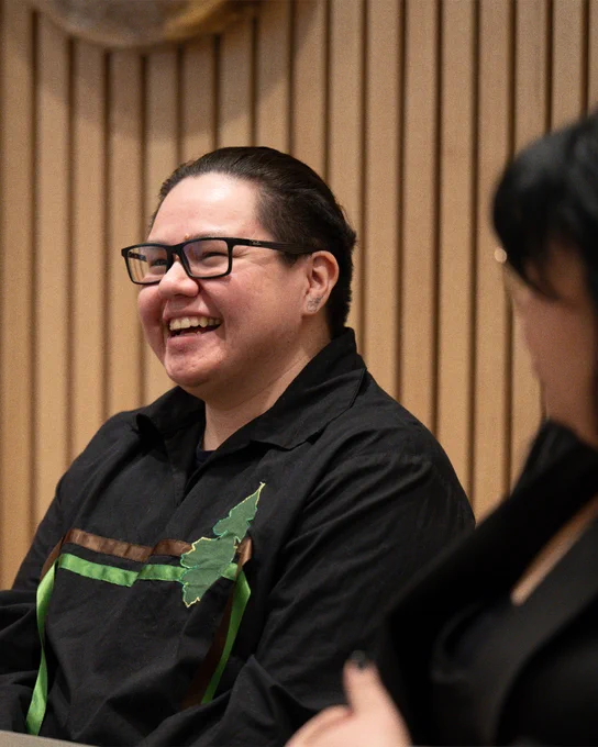 Alt text: A smiling participant wearing glasses and a black shirt with green ribbon accents engages in conversation during an information session, seated in front of a wood-panelled wall.
