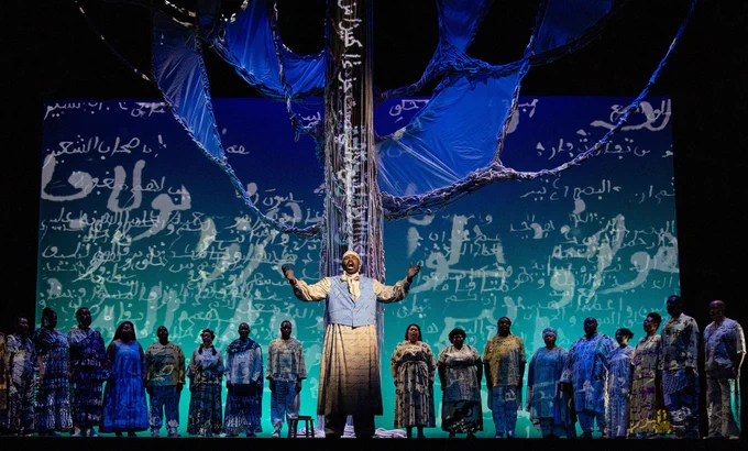 In May of 2023, Boston Lyric Opera presented Omar, the story of an Islamic scholar sold into the trans-Atlantic slave trade. A male performer stands center stage with his arms raised in celebration, wearing a light blue shirt and cream-colored pants. He is surrounded by an ensemble of performers dressed in patterned clothing in shades of blue, teal, and earth tones. Behind them is a large backdrop featuring Arabic calligraphy in white text on a gradient background that transitions from deep blue to turquoise. Dramatic blue draped fabric extends overhead from a tall central ornamental structure. Turquoise stage lighting illuminates the lower portion of the stage, creating an atmospheric theatrical scene.