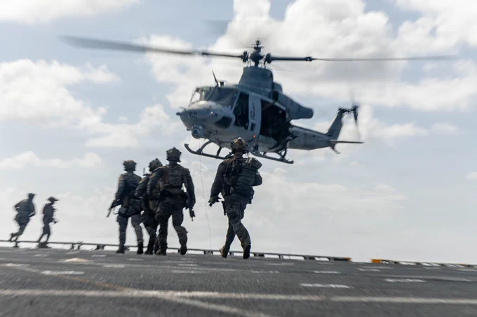 Marines move toward a hovering helicopter during deck landing and insertion operations at sea.