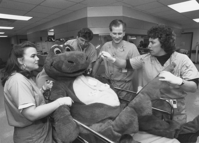 Medical staff in scrubs attend to stuffed dinosaur lying on a hospital bed in a clinical setting.