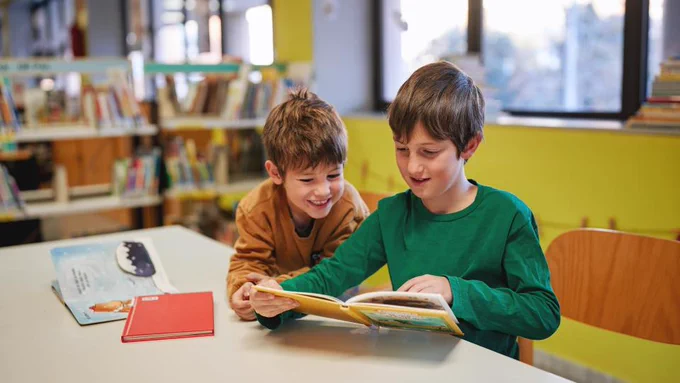 Two elementary-aged students sit at a table in a library, reading an open book together, with bookshelves in the background.